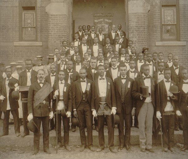 African American men posed at entrance to building, some with derbies and top hats, and banner labeled "Waiters Union" in Georgia 1899