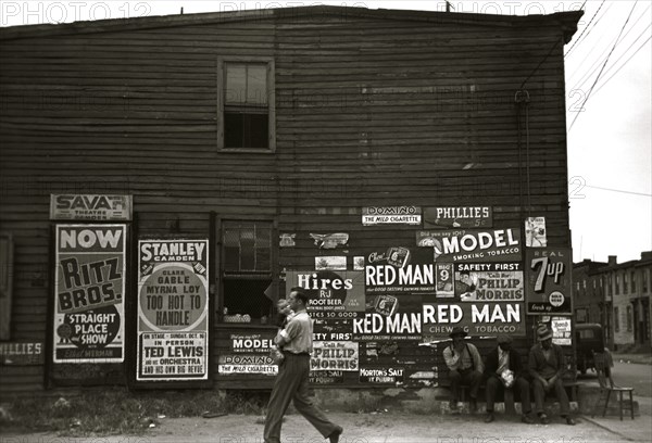 African American Men sit outside of retail establishment watching a  man walks by holding an infant 1939