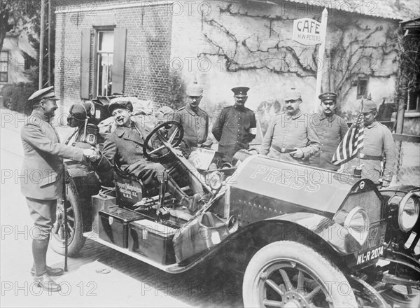 Cigar Chomping War Correspondent shakes hands with a Military person in Germany while sitting in his large limousine marked Press on the Hood in large letters; on looking military personnel are in front of a Café. 1917