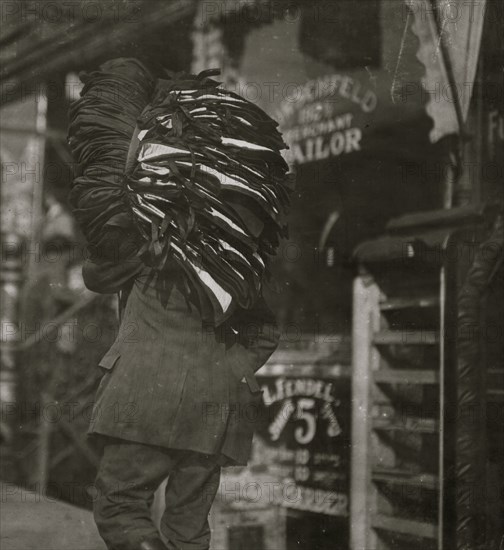 Man loaded down with heavy bundle of vests that he had been carrying for many blocks, stopping to rest, occasionally, when he would drop the bundle down onto whatever boxes or railings were available, even though they were dirty 1912
