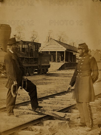 Soldier and another man standing at railroad tracks in front of locomotive 1863