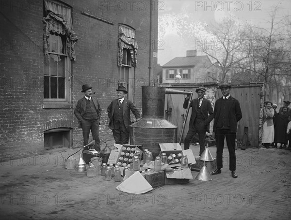 Onlookers watch as suited men stand in front of a large copper kettle still for making illegal liquor with boxes of bottles and funnels spread before them all for the manufacture of booze.