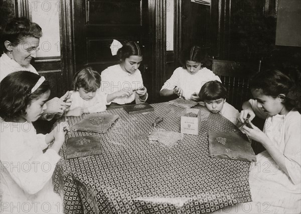 Woman and children around a table making chains for handbags 1912