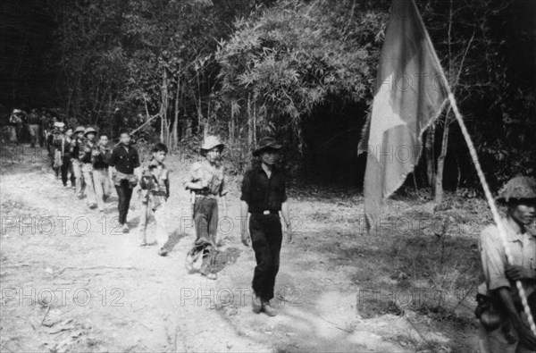 South vietnamese guerrillas file through the jungle along the ho chi minh trail, the flag with a yellow star on a red and blue background is always carried into battle, vietnam war, mid 1960s.