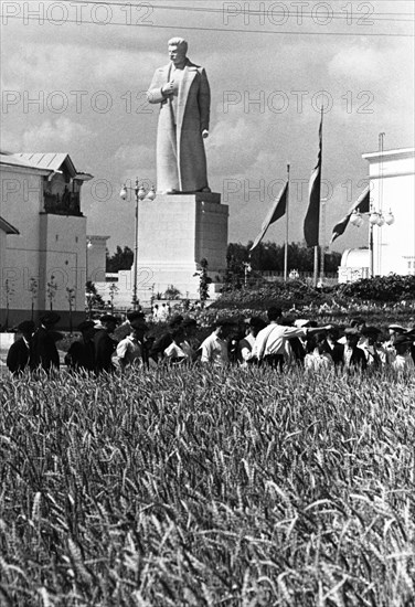 The all-union agricultural exhibition in moscow, august 1939, visitors to the exhibition examining specimens of a couch-grass / wheat hybrid (developed by member of the academy of sciences, tsitsin) at the demonstration field of the grain pavillion, in the background is a newly erected statue of stalin.