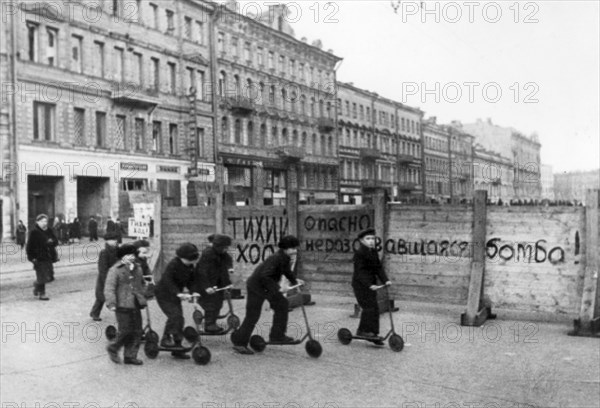 world war ll: 'drive slowly! unexploded bomb! danger!' says the notice painted on the fence, in the foreground leningrad children have organized scooter races, they have seen so much in their young lives that another unexploded bomb can neither frighten them nor detract their attention from their play.