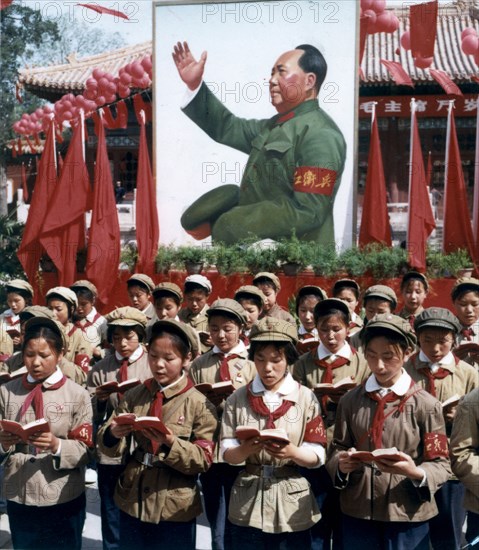 Children reading from mao's little red book at a rally with a portrait of mao in the background.
