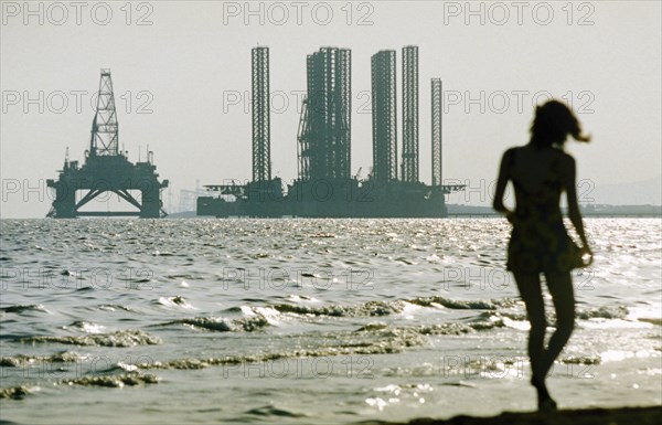 A woman in silhouette walking along shikhov beach on the shore of the caspian sea with an off-shore oil rig under construction in the background, baku, azerbaijan, october 2002.