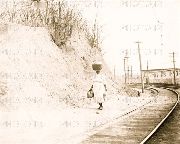 African American woman with basket on head walking near railroad tracks