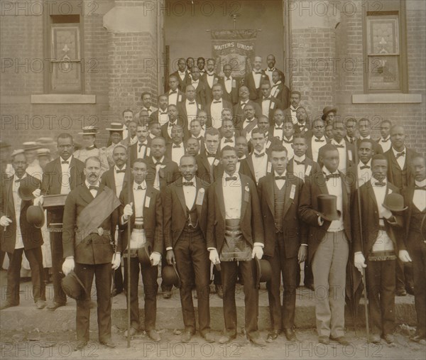 African American men posed at entrance to building, some with derbies and top hats, and banner labeled "Waiters Union" in Georgia