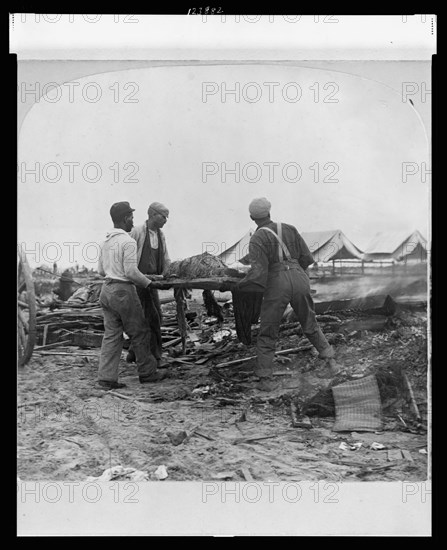 Galveston, Texas, 1900 - African-American men carrying a body on a stretcher, surrounded by wreckage of the hurricane and flood. Stereograph.