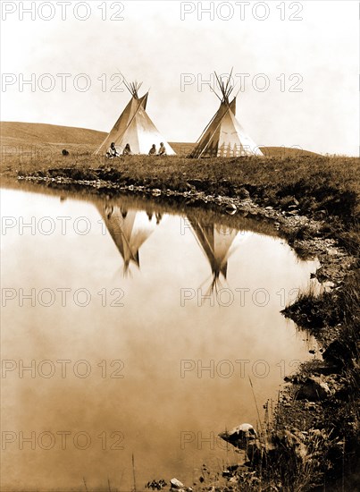 Edward S. Curits Native American Indians -  Two tepees reflected in water of pond, with four Piegan Indians seated circa 1910.