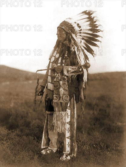 Edward S. Curtis Native American Indians - High Hawk, standing, facing left, in ceremonial dress, war bonnet and holding a coup stick circa 1907.