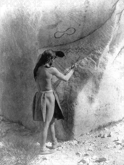 Edward S. Curtis Native American Indians - Paviotso man standing, marking side of glacial boulder that already has petroglyphs on it ca.1924.