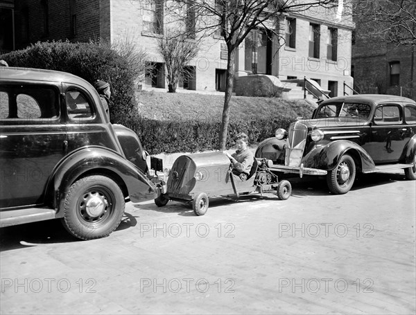 A boy occupies a parking spot in his home made go-kart circa 1937.