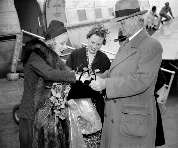 Senator William Gibbs McAdoo, of California, comes to the aid of Miss Marion Weldon, Paramount starlet, as she searches for her beauty aids before greeting the throng on her arrival at Washington Airport today. circa 1938.