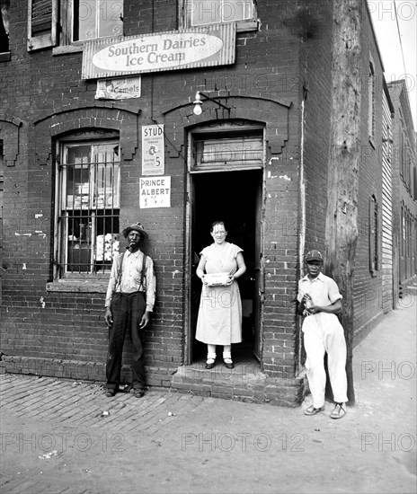 People stand outside of a small market in a slum area of Washington D.C. with Southern Dairies Ice Cream and Prince Albert ads on brick wall circa 1935 .