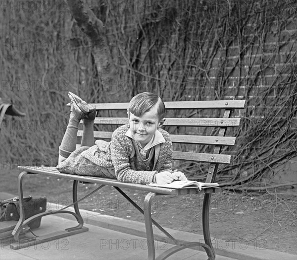 Child lying on outdoor bench with an open book ca. between 1909 and 1923