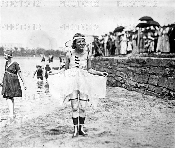 Young woman posing in bathing suit with other bathers and spectators in background ca. 1909