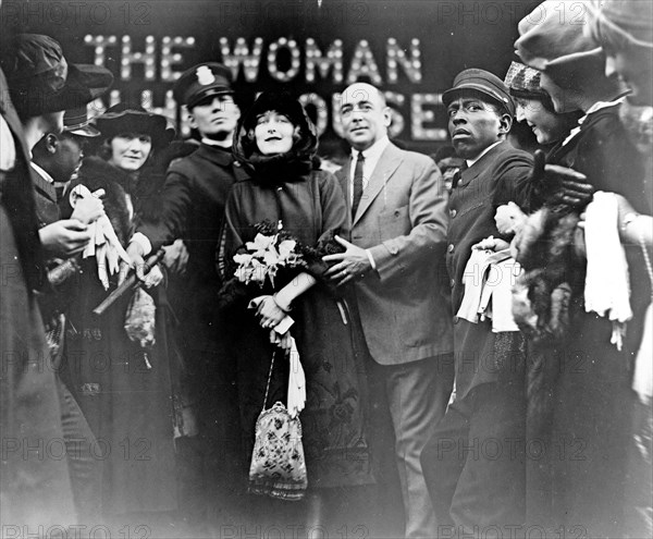 Mildred Harris Chaplin, full-length portrait, standing in crowd, facing front, guarded by a policeman (1920)