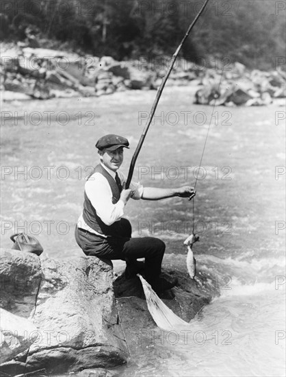 Man seated on rock, with two fish on line, by stream, possibly a river, Washington, D.C. area ca. 1909