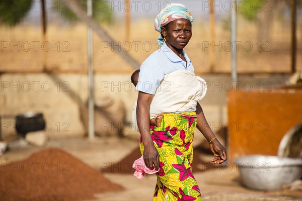 A  West Africa village woman with a baby walking towards the right in a small African village ca. 21 February 2018