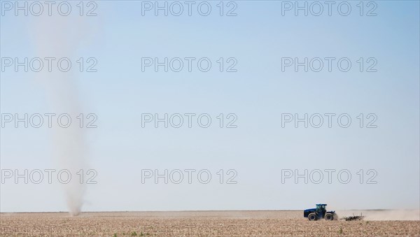 Farmer plowing a field on the Eastern Colorado plains, tractor facing left and driving toward a natural phenomena called a dust devil