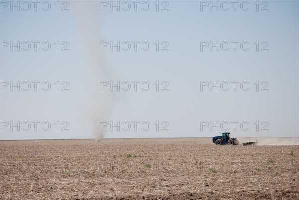 Farmer plowing a field on the Eastern Colorado plains, tractor facing left and driving toward a natural phenomena called a dust devil