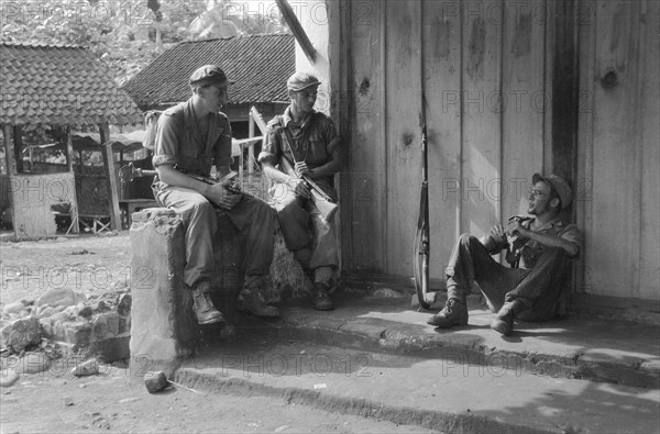 Operation Schoonschip. Three soldiers are resting in front of a building in a kampong; Date March 29, 1949; Location Indonesia, Dutch East Indies