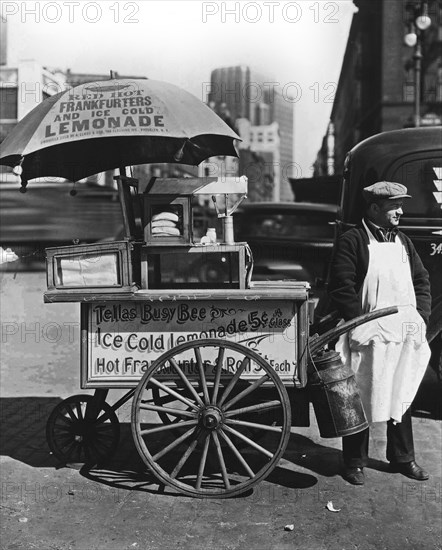 1930s New York City - Hot Dog Stand, West St. and North Moore. Vendor stands next to his Tellas Busy Bee cart, advertising 'Red Hot Frankfurters and Ice Cold Lemonade' ca. 1936