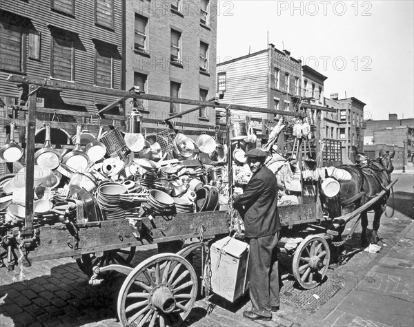 Travelling tin shop, Brooklyn. Tinker looks over his shoulder at camera while he ties box to wagon already loaded with pans, brushes, basins, etc. ca. 1936