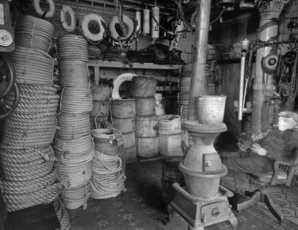 Rope store, South Street and James Slip, Manhattan. Man relaxes with cigar near pot-bellied stove, coils of ropes, life rings and other materials stacked and hung around the room ca. 1936