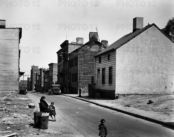 African American woman sits at street edge with two children, empty lots on either side of street, old 2 and 3 story clapboard houses further up. Talman Street, between Jay and Bridge street, Brooklyn. ca. 1936