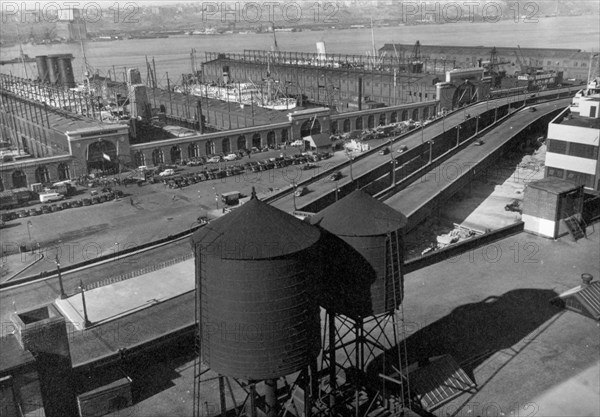Eight cars visible on Westside highway, beyond water towers in foreground, piers have entrances on street, ships visible at dock ca. 1937