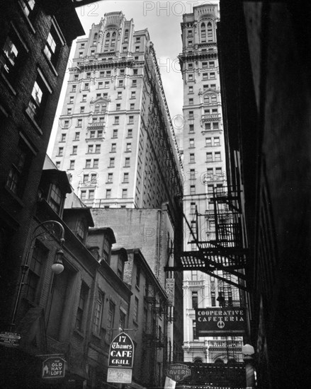 View of Thames Street, Manhattan New York City street scene. Low, older buildings, signs for bars, cooperative cafeteria, etc., street lamps and firsecapes in foreground, tall buildings in the light beyond ca. 1938