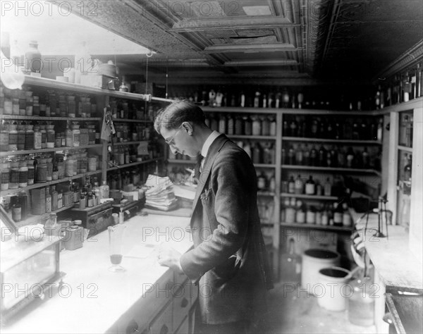 Pharmacist at People's Drug Store, No. 5, 8th and H Streets, N.E., Washington, D.C., looking at prescriptions(?) on the counter in room lined with shelves of pharmacy bottles ca. 1909-1932
