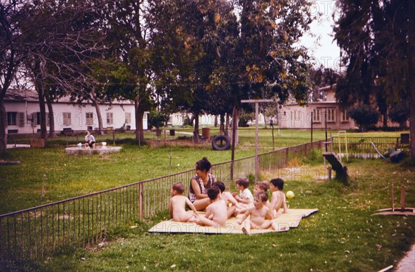 Israel April 1965: Toddlers enjoying the fresh air, wearing no clothes, with a woman caretaker on an Israeli Kibbutz