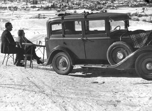 le dimanche à la montagne d'une famille dans les années 1930 : la voiture fiat, les tables et chaises pliantes pour le petit-déjeuner, le plaid et les skis, 1930