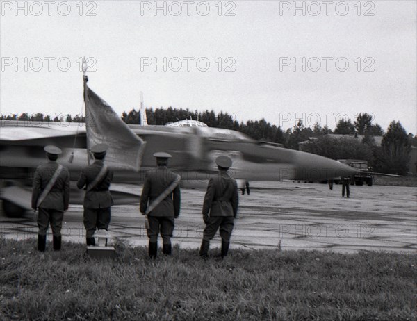 Four officers with regimental banner stand against the MiG-23 taxiing out for takeoff, air base Kubinka, USSR.