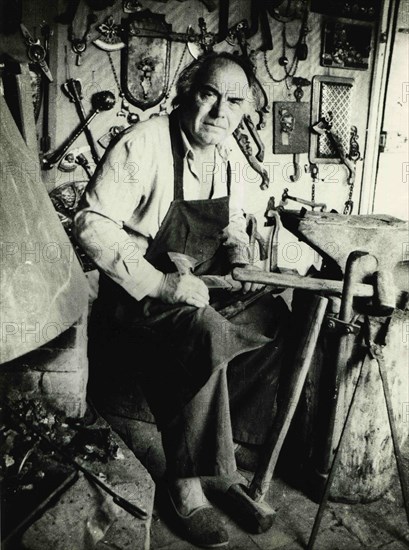 A blacksmith sits near an anvil in his own forge.