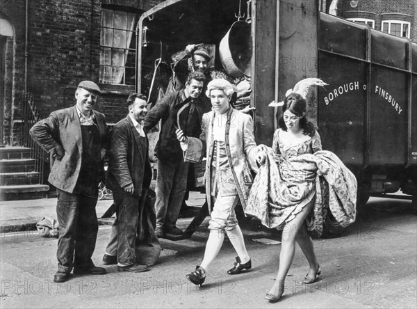 A couple of masked young people pass in front of a group of workers, london, england, uk. 1967