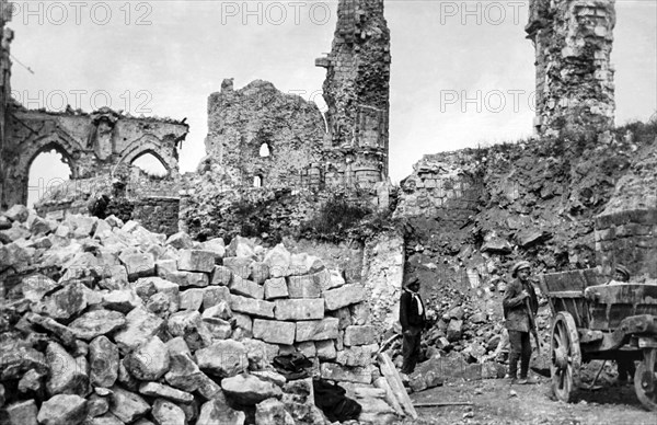 Ruins of buildings and a pile of rubble and stone blocks with workers standing by a wooden cart.