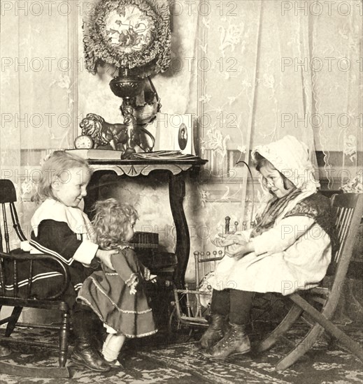 Two little girls playing with a doll, one dressed as a mummy and the other as grandma beconing the doll to come to her.