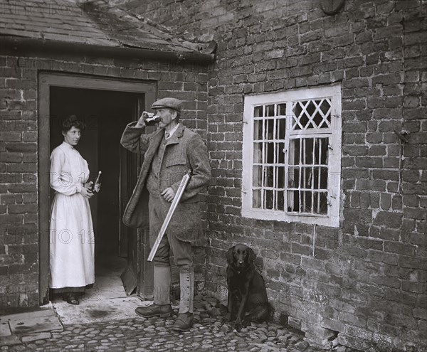A man with a gun drinking beer and a lady in the doorway of an old cottage holding bottle and waiting for glass.