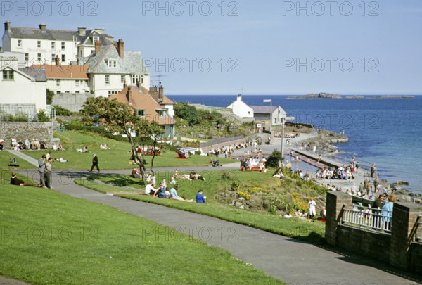 People sittng on grass by the coast enjoying sunshine at the seaside resort of Portrush, County Antrim, Northern Ireland, UK 1960s.