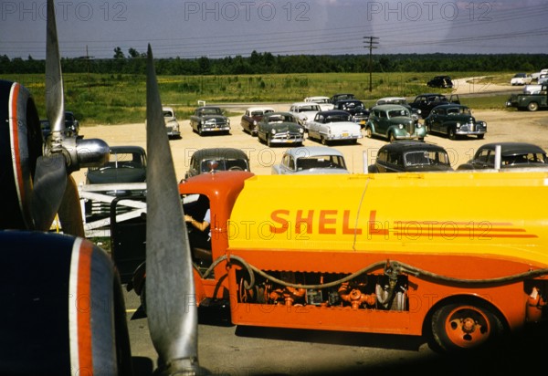 Shell oil fuel tanker vehicle viewed through airplane window with cars parked beyond, USA c 1953.