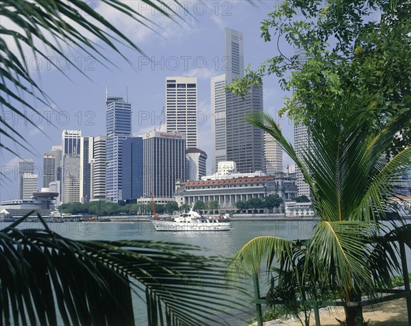 SINGAPORE, Singapore City, City skyscrapers and waterfront seen through palm leaves wth a white cruiser sailing past