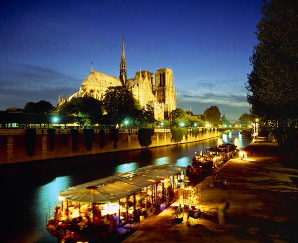 FRANCE, Ile de France, Paris, Notre Dame floodlit at night with restaurant and houseboat lit up on the River Seine in foreground.