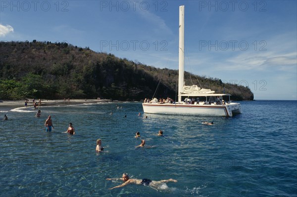 WEST INDIES, St Lucia, Transport, Catamaran tour.  Tourists swimming from moored boat with beach and tree covered headland behind.