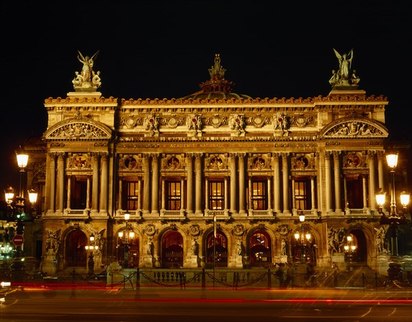 FRANCE, Ile de France, Paris, Opera frontage floodlit at night against black sky with light trails from passing traffic in the foreground.